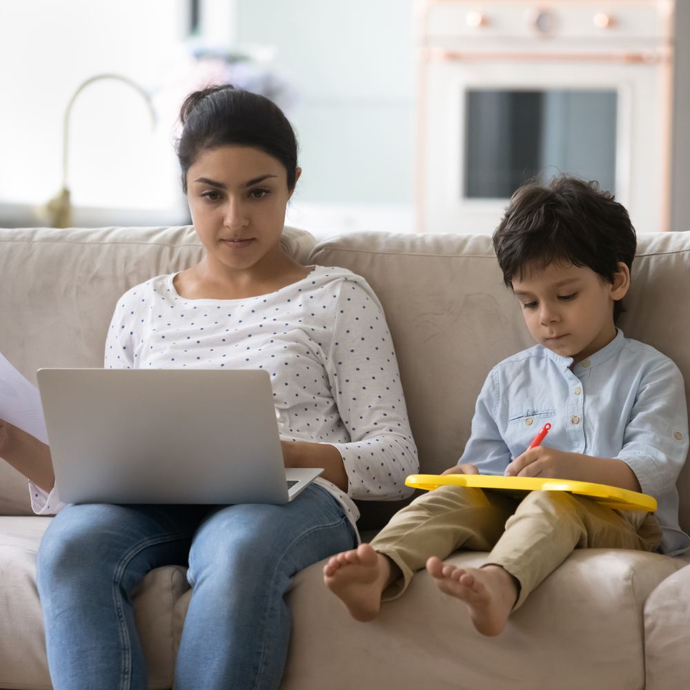 Mutter erledigt Unterlagen am Laptop, ihr Sohn malt neben ihr auf dem Sofa