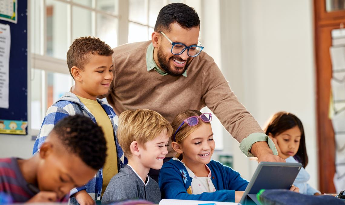 Ein Betreuer schaut lachend mit einer Gruppe Grundschulkinder auf ein Tablet